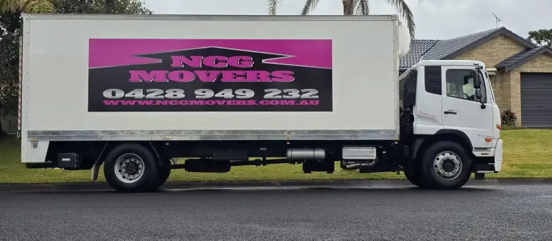 An NCG Movers truck side on in a suburban street, showing the full pink and black livery