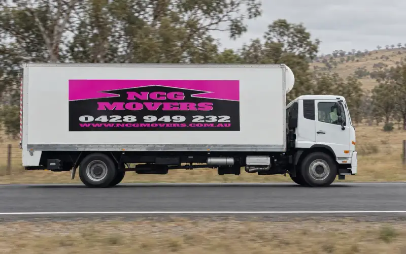 A white NCG Movers truck driving along a rural highway past gum trees and dry paddocks